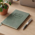 A green notebook with an embossed logo and a silver pen on a wooden desk, alongside a potted plant.