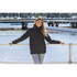 A woman wearing a black puffer jacket stands by the water, with a marina and bridge in the background.