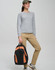 A woman wearing a grey top and khaki pants holds a black and orange Iconic Promo Backpack.