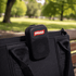 A black portable speaker with a logo, attached to a bag, set against a green park background.