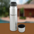 A drink bottle with a silver stainless steel finish, featuring a black lid and a detachable black cup. It has a logo.