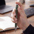 A green metal pen held in a hand, featuring a branded logo, with a blurred workspace in the background.