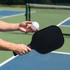 A black pickleball paddle held in a hand, alongside a white perforated pickleball, on a court with nets in the background.