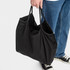 A black nylon shopper bag with two handles, being held by a person wearing jeans and sneakers.