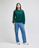 A green crewneck sweatshirt is worn with light blue jeans by a standing model in a neutral background. The sweatshirt has a logo.