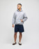 A man stands wearing a grey pullover hooded sweatshirt and navy shorts. The sweatshirt features a logo.