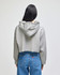 A cropped grey hoodie with a hood, viewed from the back, paired with blue jeans. It features a branding logo.
