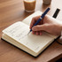A diary with cream-coloured pages and a pen resting on a wooden table, alongside a coffee cup.