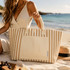 A striped beige and cream tote bag with handles, sitting on a beach next to a woman in a sundress.
