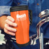 An orange travel mug with a black lid and handle, featuring a logo, held by a person next to a bicycle.