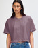 A women's cropped boxy T-shirt in a muted purple shade, featuring a logo. The model is styled with jeans.