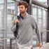 A man wearing a light grey half-zip sweatshirt, carrying a shoulder bag, walking in a modern building.