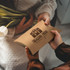 A large pillow box in brown cardboard, featuring a logo design, held by two hands with a bouquet of tulips.