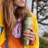 A drink bottle in green with a botanical design, held by a person wearing a yellow jacket near trees.