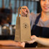 A small, brown paper bag with flat handles and a logo, being held by a person in an apron.