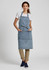A denim apron in blue, worn by a model over a white t-shirt, featuring pockets and a tied waist.