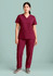 A woman stands wearing burgundy straight leg scrub pants and a matching short-sleeve top. The outfit has two pockets.