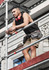 A man wearing a black sleeveless tee, shorts, and a tool belt stands on scaffolding, with headphones around his neck.