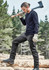 A man in black work pants and a long-sleeve top, holding an axe while standing on logs in a forested area.