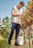 A man in a beige long sleeve outdoor shirt and navy pants is tending to grapevines in a vineyard.