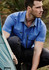 A short-sleeve, blue button-up shirt with one chest pocket and logo, worn by a man outdoors near a tent.