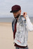 A man at the beach wears a burgundy cap and a denim jacket with graphic details. The ocean is in the background.