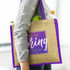 A jute tote bag in natural and purple, carried with a drink bottle and an apple visible inside.