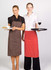 Two women wearing striped full bib aprons, one in brown and the other in red, holding food and drinks.