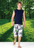 A man wearing a navy muscle T-shirt and floral shorts, standing on a grassy area with trees in the background.