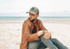 A man with a cap sits on the beach, wearing a brown jacket and grey jeans. The cap is olive green with a logo.