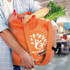 An orange tote bag filled with vegetables, including celery, capsicum, and a large root vegetable. It has a logo.