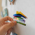 A hand holds a colourful house-shaped sticker on a light surface, featuring a logo and vibrant rainbow design.