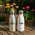 A drink bottle set featuring a silver stainless steel bottle and a white bottle, both with logos, amid blooming flowers.
