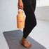A drink bottle in a peach colour held by a person in black leggings, set on a yoga mat in a bright space.