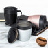 A set of three insulated coffee mugs in black, silver, and rose gold with lids, placed on a marble surface.