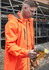 A man wearing an orange unisex packable rain jacket, focused on using a power tool on a construction site.