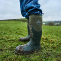 Muck Boots MUCK Boots Chore 2k Mens Classic Stable Farm Tall Wellington Wellies, green weatherproof rubber boots covered in mud, worn on a grassy farm field.