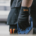Scruffs Trade Work Men's Padded Palm Gloves, close-up of a person's hand wearing the black glove with orange logo, gripping a hand saw for construction or carpentry work.