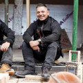 Scruffs Trade Flex Softshell Mens Zip Up Work Warm Jacket Coat, a man wearing black work jacket and pants, sitting on the floor at a construction site, smiling and looking at the camera.