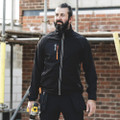 Scruffs Trade Flex Softshell Mens Zip Up Work Warm Jacket Coat, a man wearing a black softshell jacket with orange zipper details, standing at a construction site with a brick wall in the background.