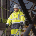Scruffs High Visibility Mens Hi-Viz Waterproof Zip Up Jacket Coat, a man wearing a high-visibility yellow waterproof jacket with reflective stripes and a white safety helmet at a construction site.