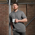 Scruffs Tech Polo Mens Work Cotton Short Sleeve Professional Polo, man holding a white mug in a casual pose standing against an industrial brick wall background.