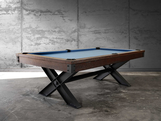Three-quarter angled view of the Nixon Portland Pool Table. The rustic wood cradle pairs with sleek industrial steel legs, while the blue Teflon-coated Championship felt adds a pop of color against the neutral game room backdrop.