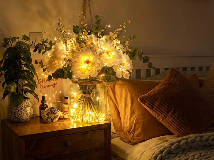 A glowing jar of fairy-light blooms on a bedside table beside a bed with a knit throw