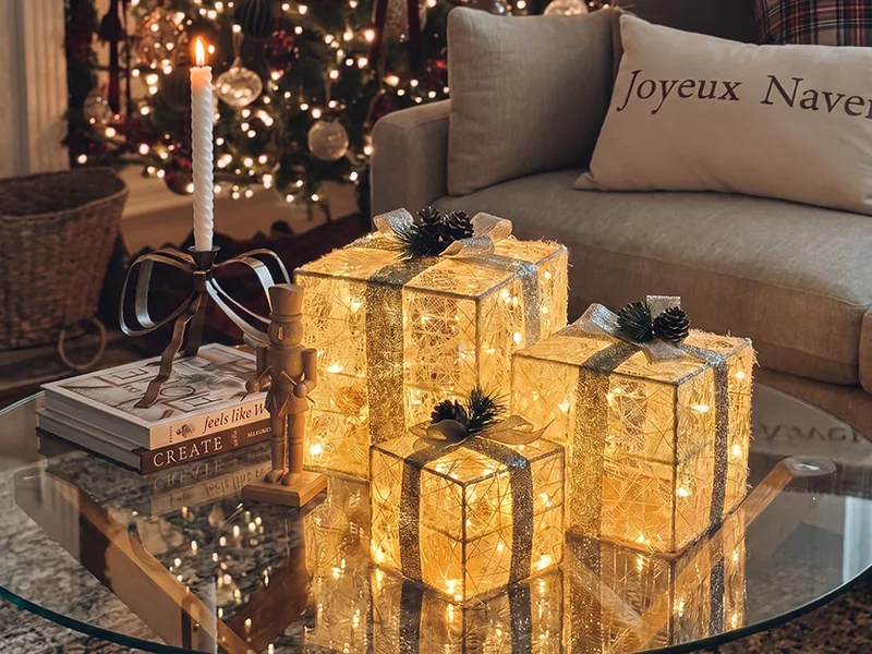 Three glowing gift-box lights stacked on a glass coffee table in front of a Christmas tree