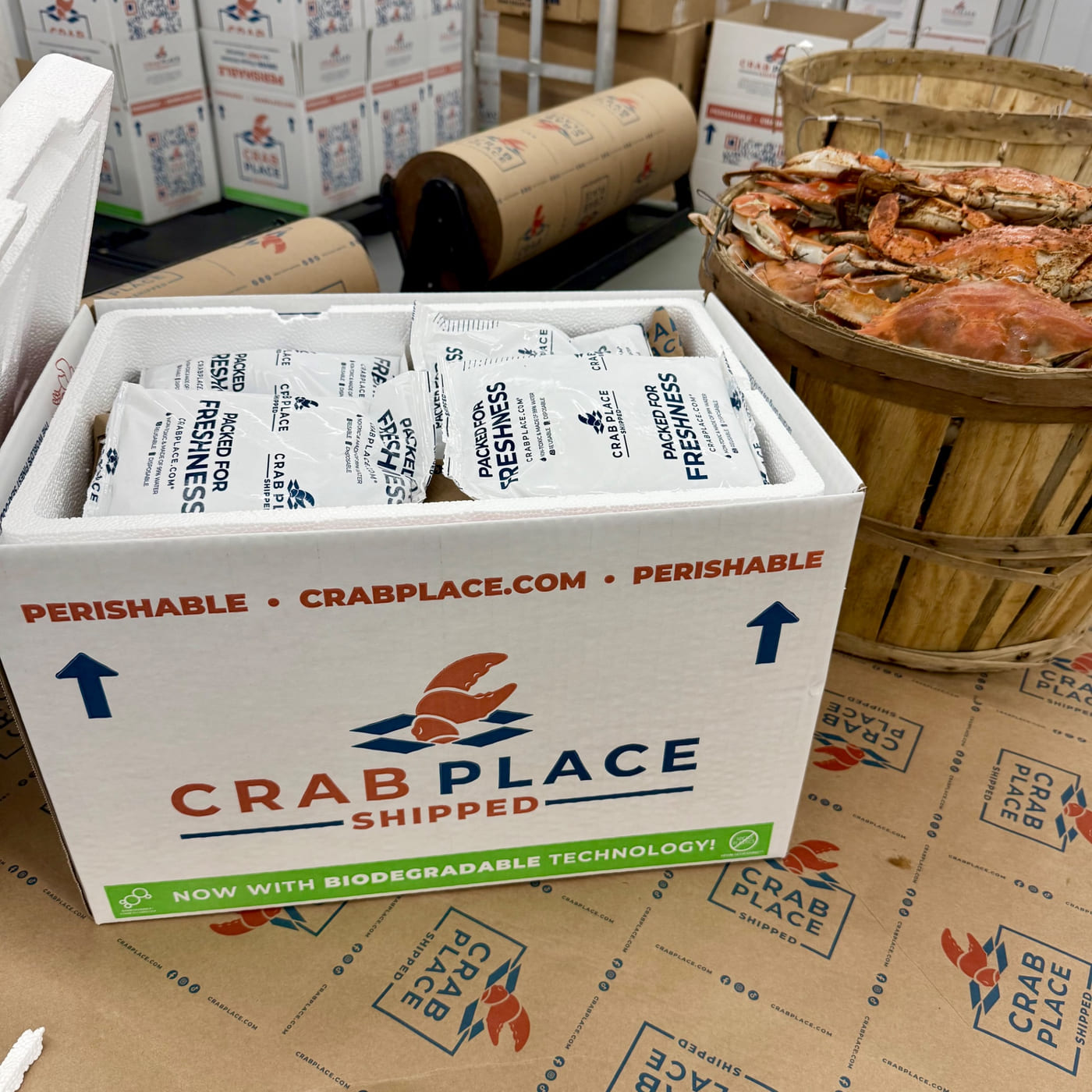 A Crab Place shipping box packed tight with Packed for Freshness gel packs, beside a bushel of steamed Maryland blue crabs