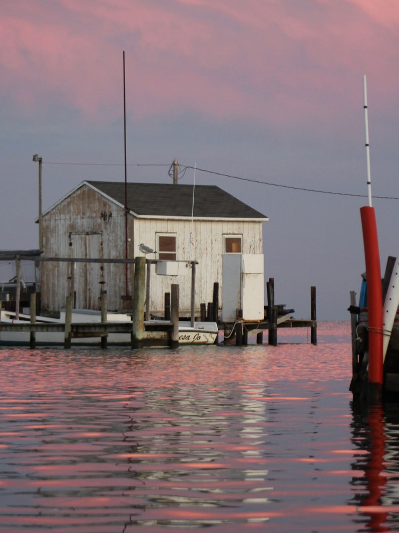 A weathered crab shanty on pilings over the water at dusk on Tangier Island, with a workboat tied up alongside.