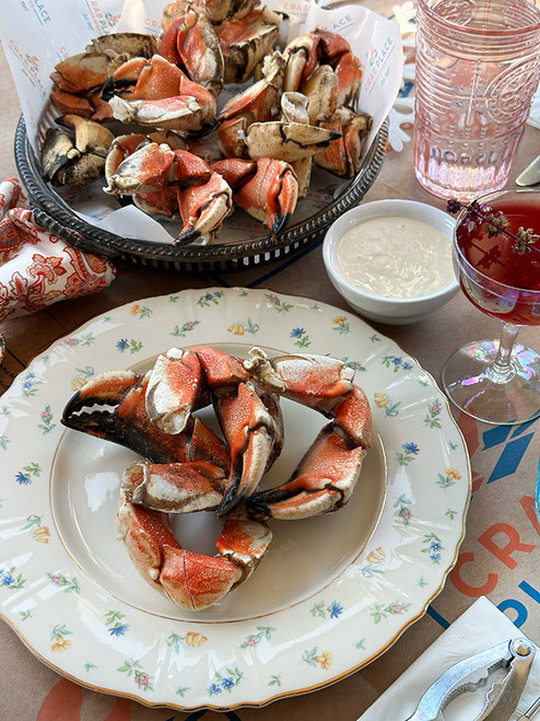 Scored Jonah crab claws on a floral plate and silver tray, served with a dipping sauce, a pink cocktail, and a crab cracker.