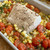 Baked Alaskan black cod fillet with cracked pepper on roasted cherry tomatoes, sweet corn, and parsley in a metal pan.
