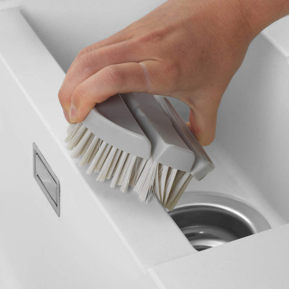 Grey handled cleaning brush being used to scrub a white kitchen sink edge